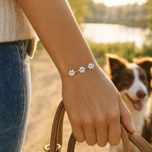 Main tenant une laisse en cuir au bord de l'eau, bracelet Câline argent à trois pattes, un border collie à côté