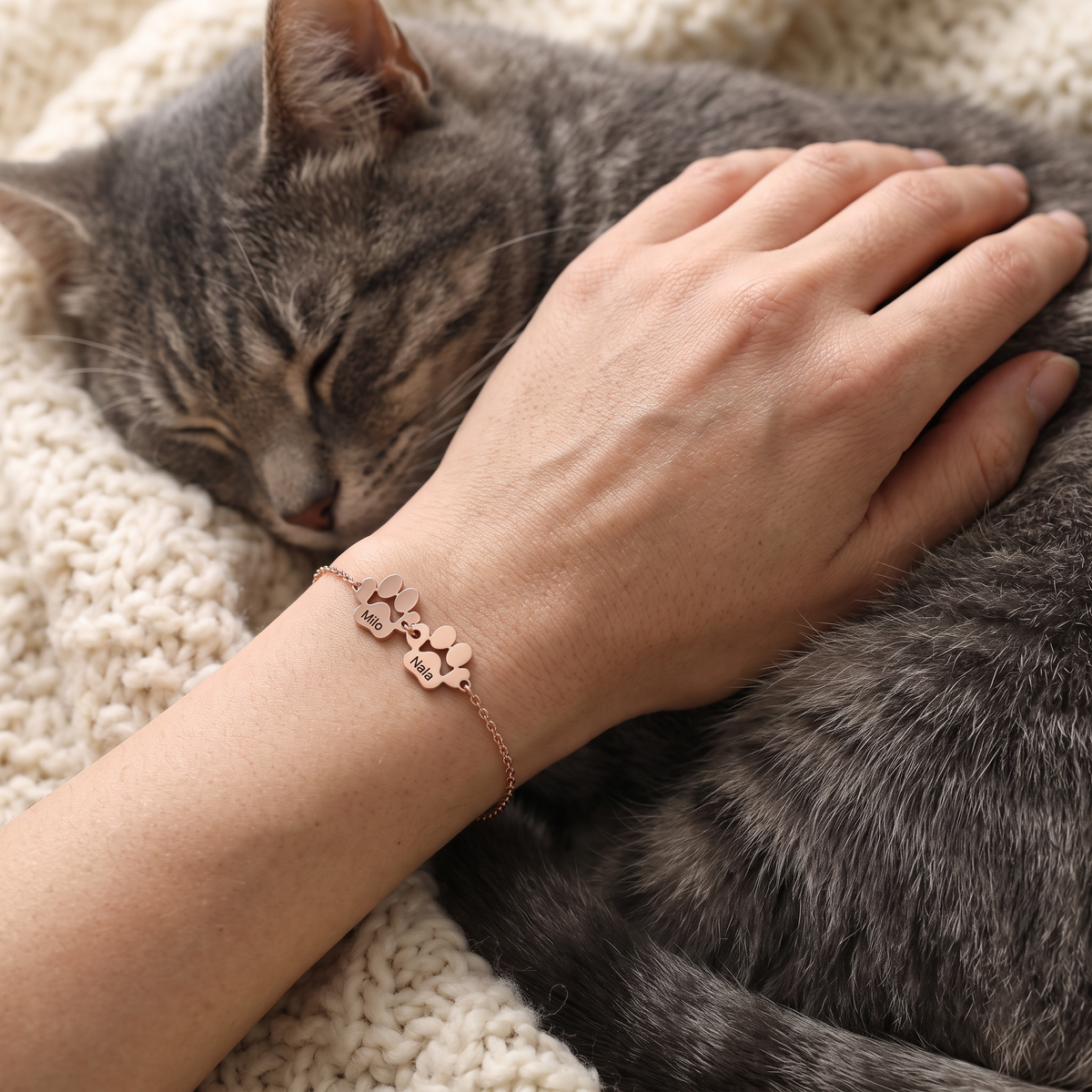 Main portant un bracelet Câline or rose à 2 pattes gravées Milo et Nala, posée sur un chat endormi